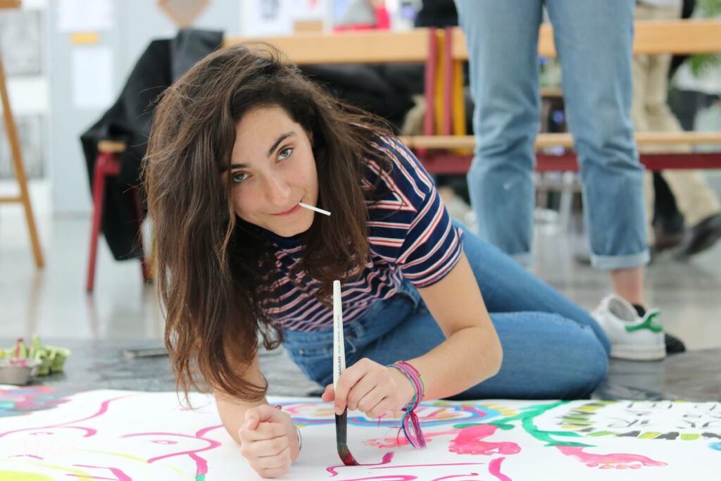 femme qui est part terre et fait de la peinture sur une grande affiche sur le sol, et regarde la caméra
