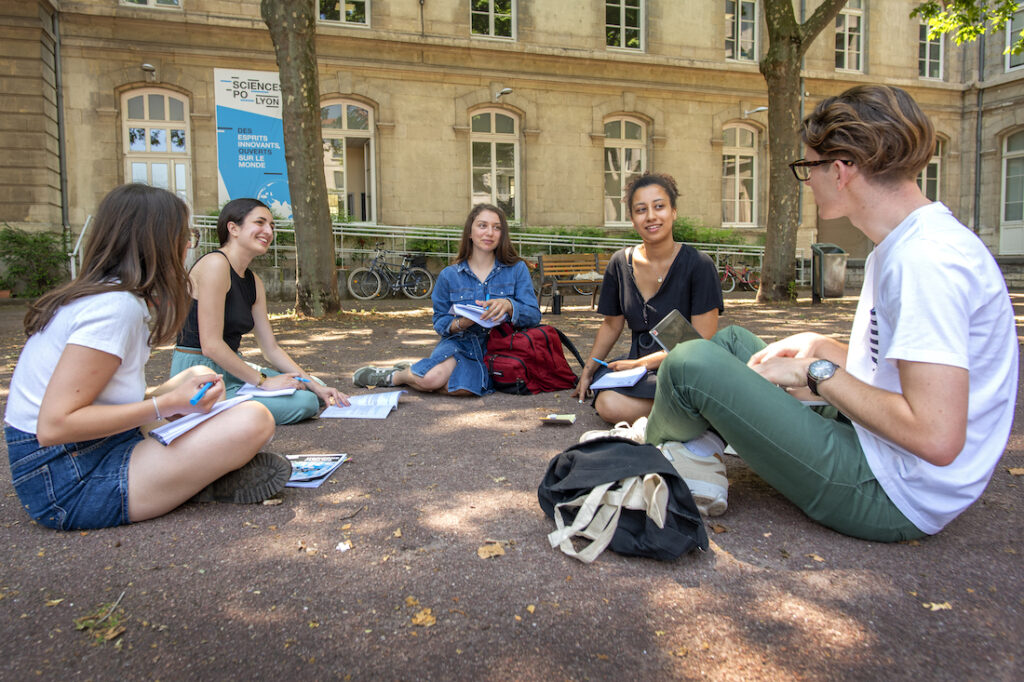 5 élèves assis par terre, dans la cour, avec un ordinateurs et des notes