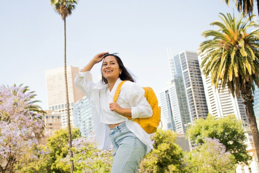Femme avec un grand sourire, qui se tient les lunettes du soleil sur sa tête, un sac à dos jaune sur le dos. Des palmiers et des arbres à fleurs violettes en arrière plan