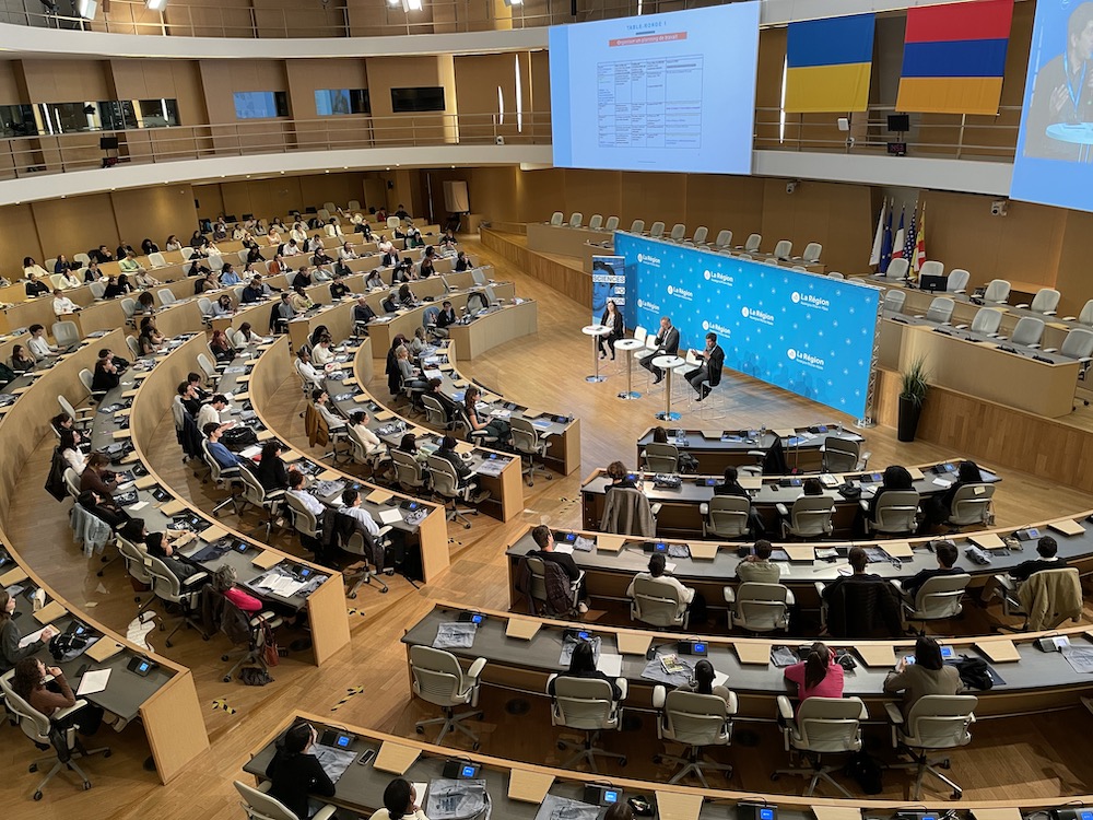 Amphithéâtre rempli de personnes regardant trois personnes qui parlent, vue du haut