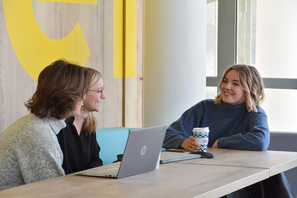 3 étudiantes autour d'une table qui sourient, dont l'une avec un café à la main