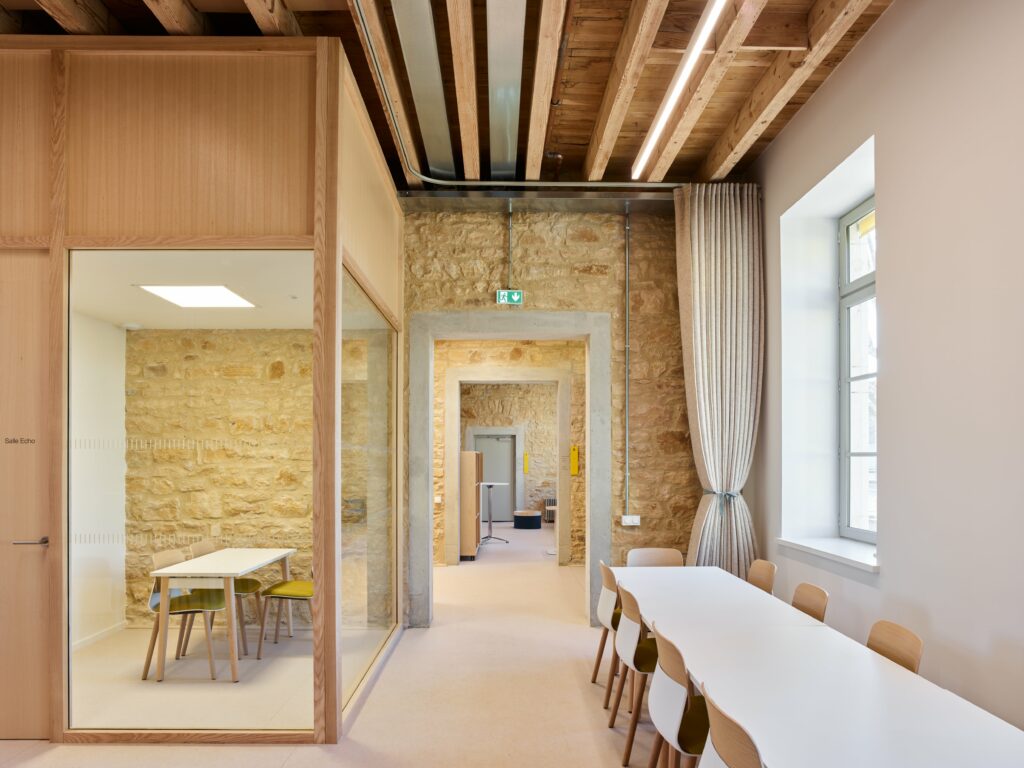 Pièce avec une grande table sur la droite, vers une fenêtre. A gauche, une petite salle avec les murs en vitre qui permet de voir dedans, avec une petite table et trois chaises.