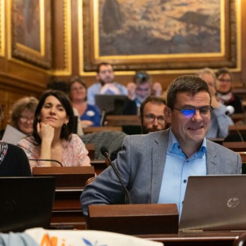 Un homme qui sourit, assis avec un ordinateur devant lui, à côté d'une femme. En arrière plan, on d'autres personnes assis sont floutés, dans un amphithéâtre