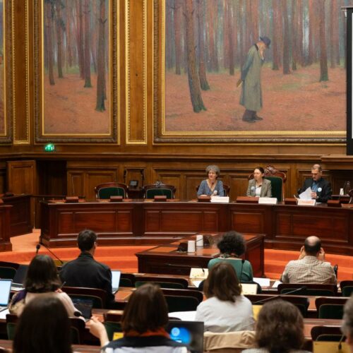 2 femmes et 1 homme devant une audience dans un grande salle avec des tables en bois et des grands tableaux