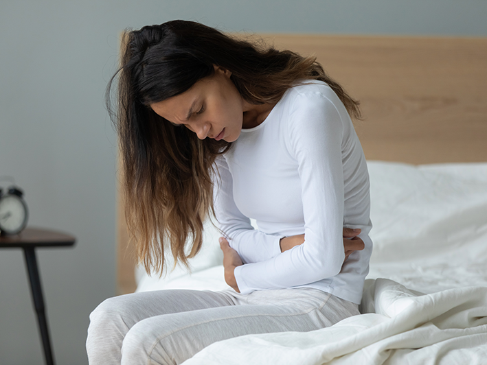 Femme habillé en blanc, assis sur un lit blanc, qui met ses mains sur son ventre avec une expression de douleur