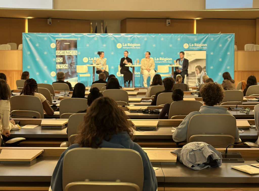 Au premier plan, des personnes de dos assis dans un amphithéâtre qui regarde 4 personnes face à eux qui parlent au micro