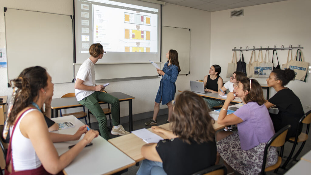 2 élèves qui font une présentation en regardant le vidéoprojecteur. Plusieurs femmes les écoutent sur des tables en forme de U