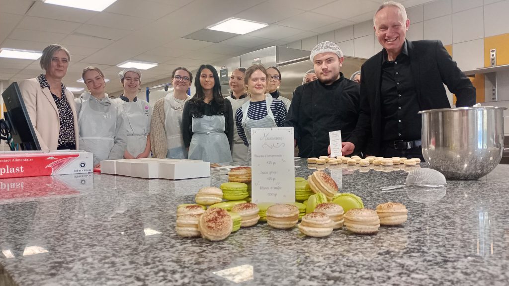 Photo d'un groupe de personnes dans une cuisine. En premier plan, des macarons et une feuille décrivant la recette